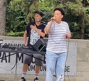 An outdoor stage with 2 singers. One is singing in front of a DJ setup, the other is holding a mic. 