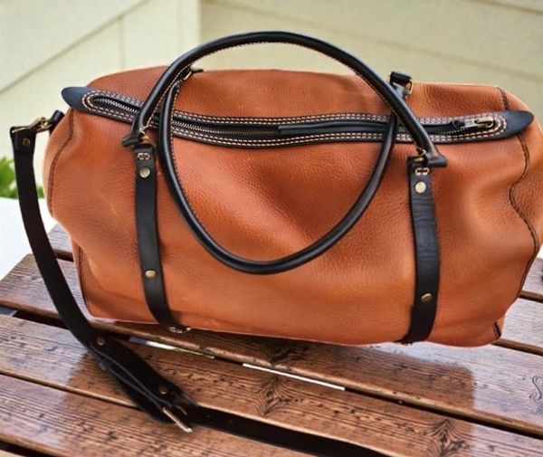 Brown leather duffel bag with black handles on a wooden surface.