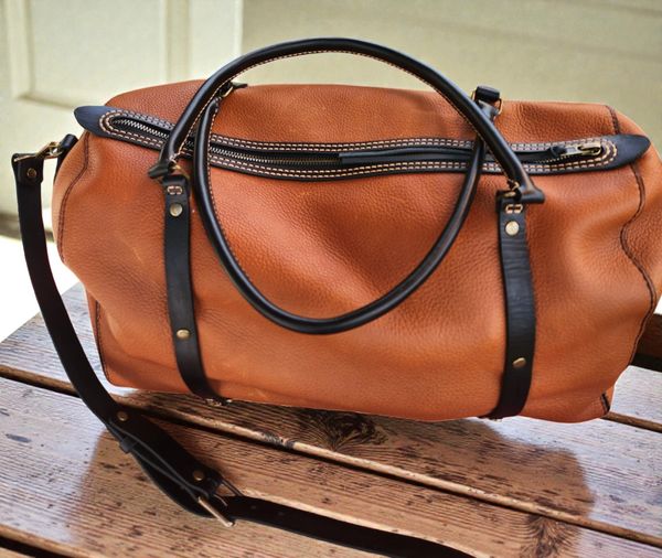 A stylish brown leather duffel bag with black handles and shoulder strap on a wooden surface.
