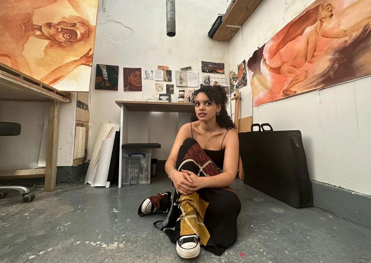 Natalie Charles, artist, sitting on the floor of her studio with artwork hanging on the walls.