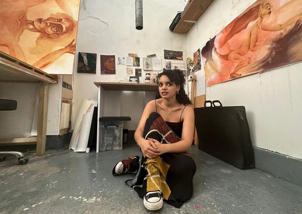 Natalie Charles, artist, sitting on the floor of her studio with artwork hanging on the walls.