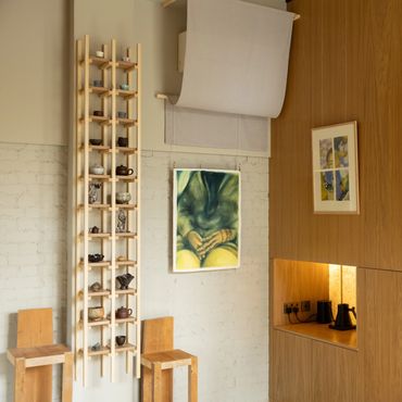 Minimalist room with wooden chairs, a tall shelf of teapots, and framed art on white and wood walls.