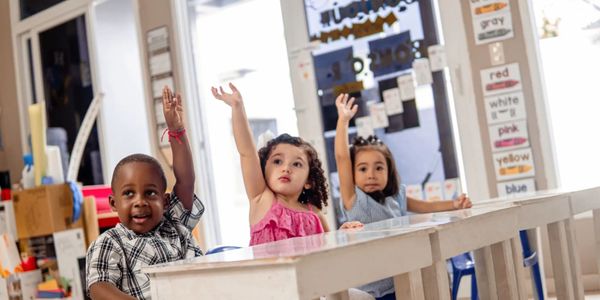 Three young children sitting at desks with hands raised in a classroom.