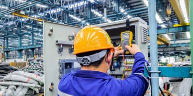 Technician in a yellow helmet checking electrical panel in a factory.