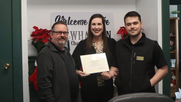 Three people smiling, one holding a certificate in an office setting with festive decorations.