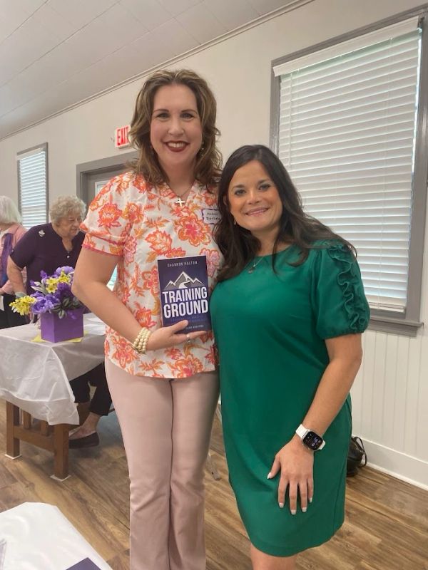 Two women smiling, one holding a book titled 'Training Ground.'