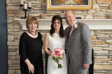 Bride in white dress with two companions posing indoors by a stone fireplace.