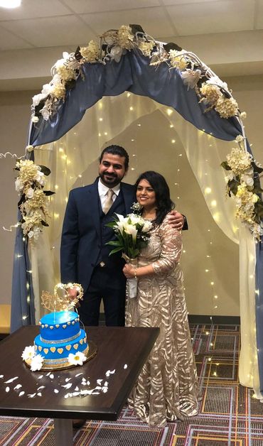 Couple standing under a floral arch with a blue cake on the table.