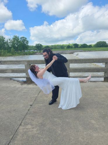 Groom dips bride in white dress on a bridge by the water under a bright sky.