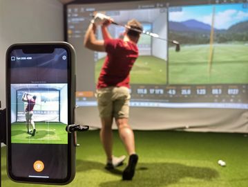 Man practicing golf swing indoors with a golf simulator and phone recording.