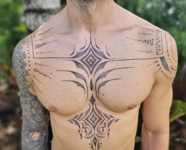 Intricate tribal tattoos cover the chest and arms of a shirtless man outdoors.