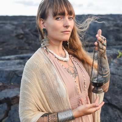 Woman holding incense burner, adorned with jewelry and tattoos, standing outdoors.