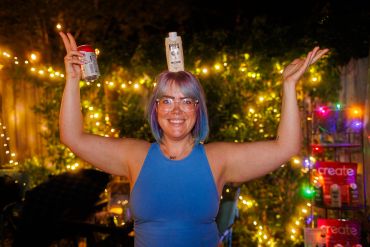Smiling woman with purple hair holding bottles, surrounded by festive lights outdoors.