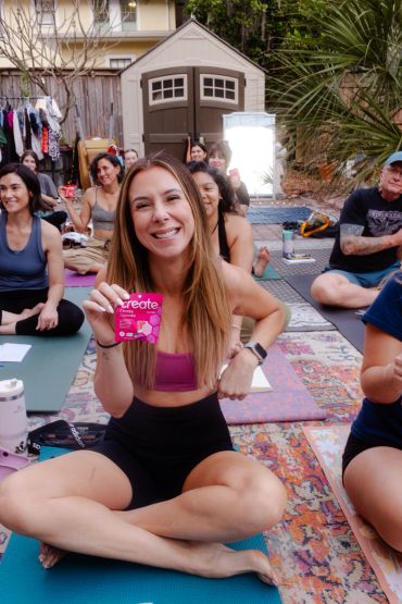 Smiling woman in workout clothes holds a pink product while sitting cross-legged on a yoga mat in a group class.