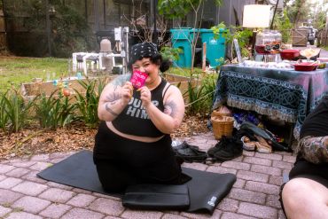 A woman sitting on a mat outdoors, eating a snack with a smile.