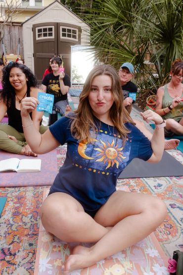 A woman flexes her arms and holds a card, surrounded by people sitting on mats outdoors.