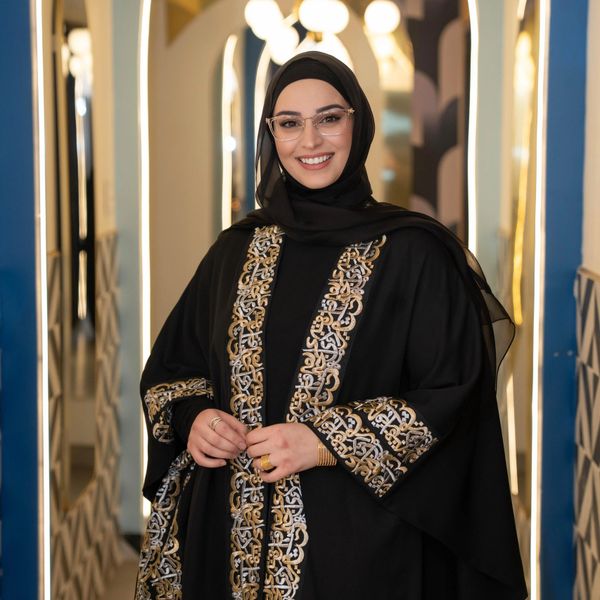 Smiling woman wearing a black abaya with intricate gold embroidery and glasses.