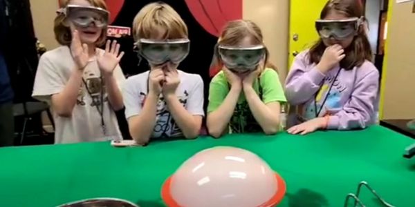 Four kids in goggles watch a large bubble on a table with excitement.