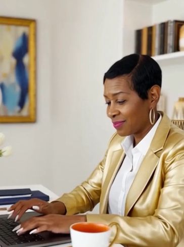 Woman in gold jacket working on laptop with tea cup nearby.