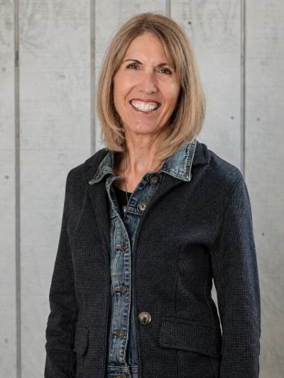 Smiling woman in layered denim and blazer against a wooden background.