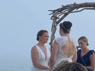 Two women exchanging vows at an outdoor wedding ceremony by the water.