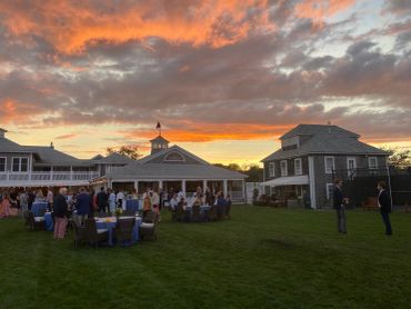 Outdoor evening gathering with tables and people under a colorful sunset sky.