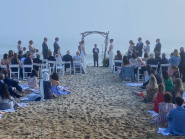 A beach wedding ceremony with guests seated on chairs and blankets.