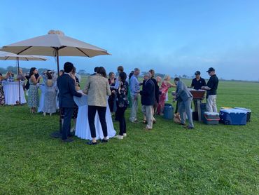 People socializing outdoors around tables and coolers on a grassy field.