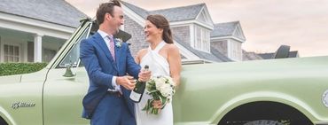 Newlyweds celebrating with champagne by a vintage green car outside a house.