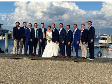 Bride with groom and groomsmen posing on dock by water under a cloudy sky.