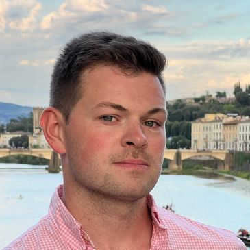 A man in a pink shirt posing near a river with buildings in the background.
