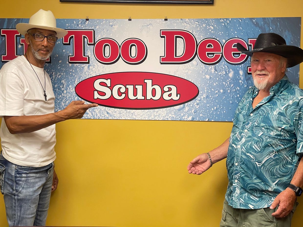 Two men in hats pose in front of a 'Scuba' sign on a yellow wall.
