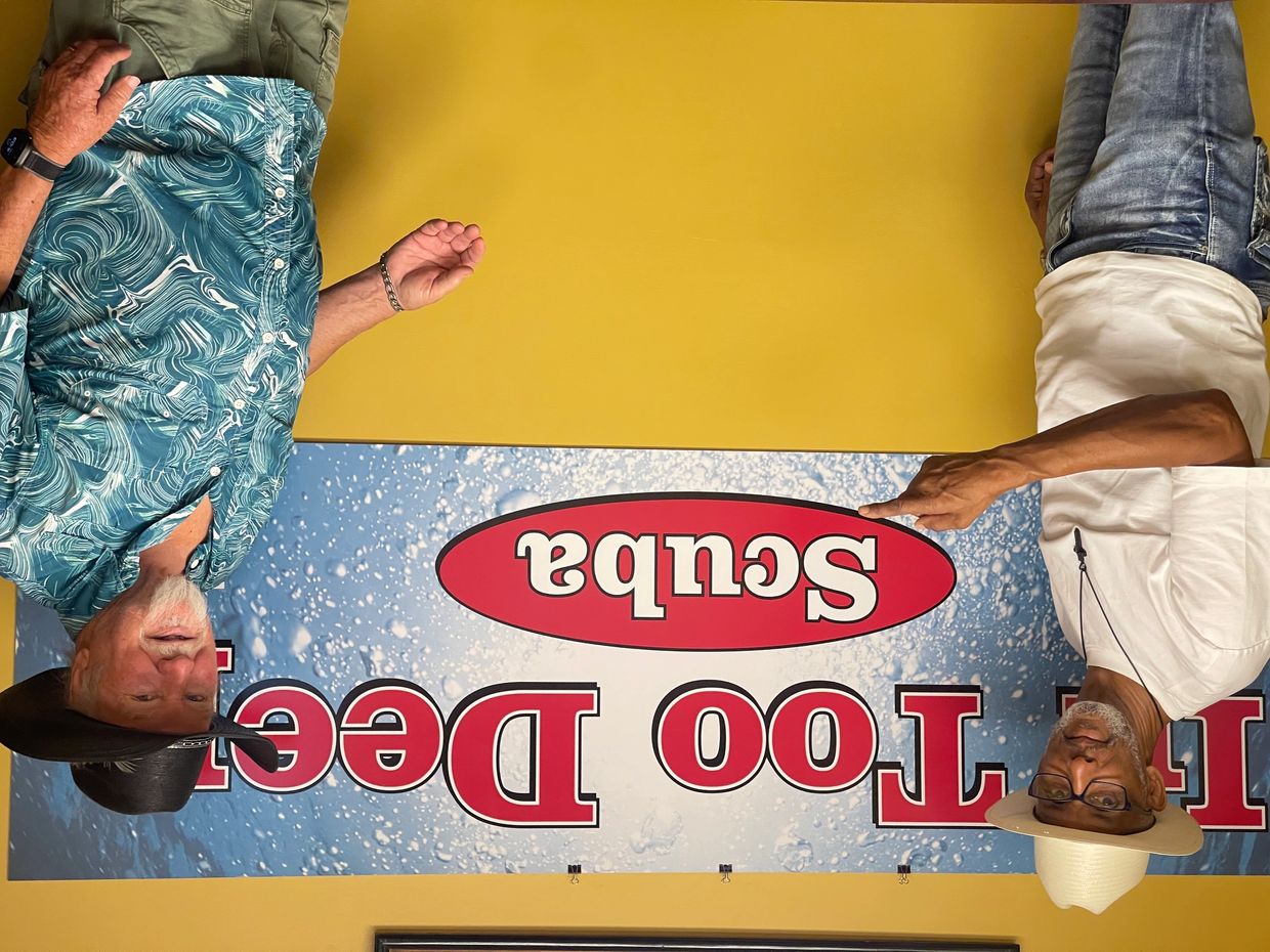 Two men in hats pose in front of a 'Scuba' sign on a yellow wall.