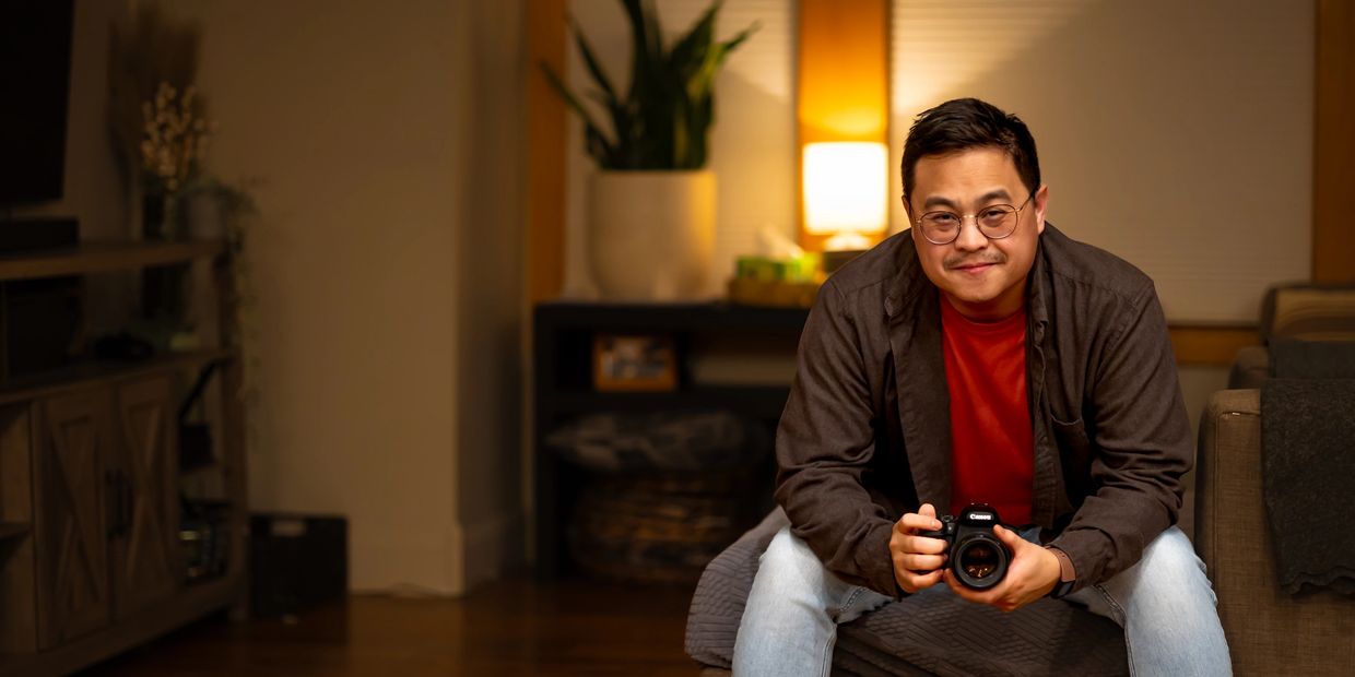 Man sitting indoors holding a Canon camera, smiling at the camera.