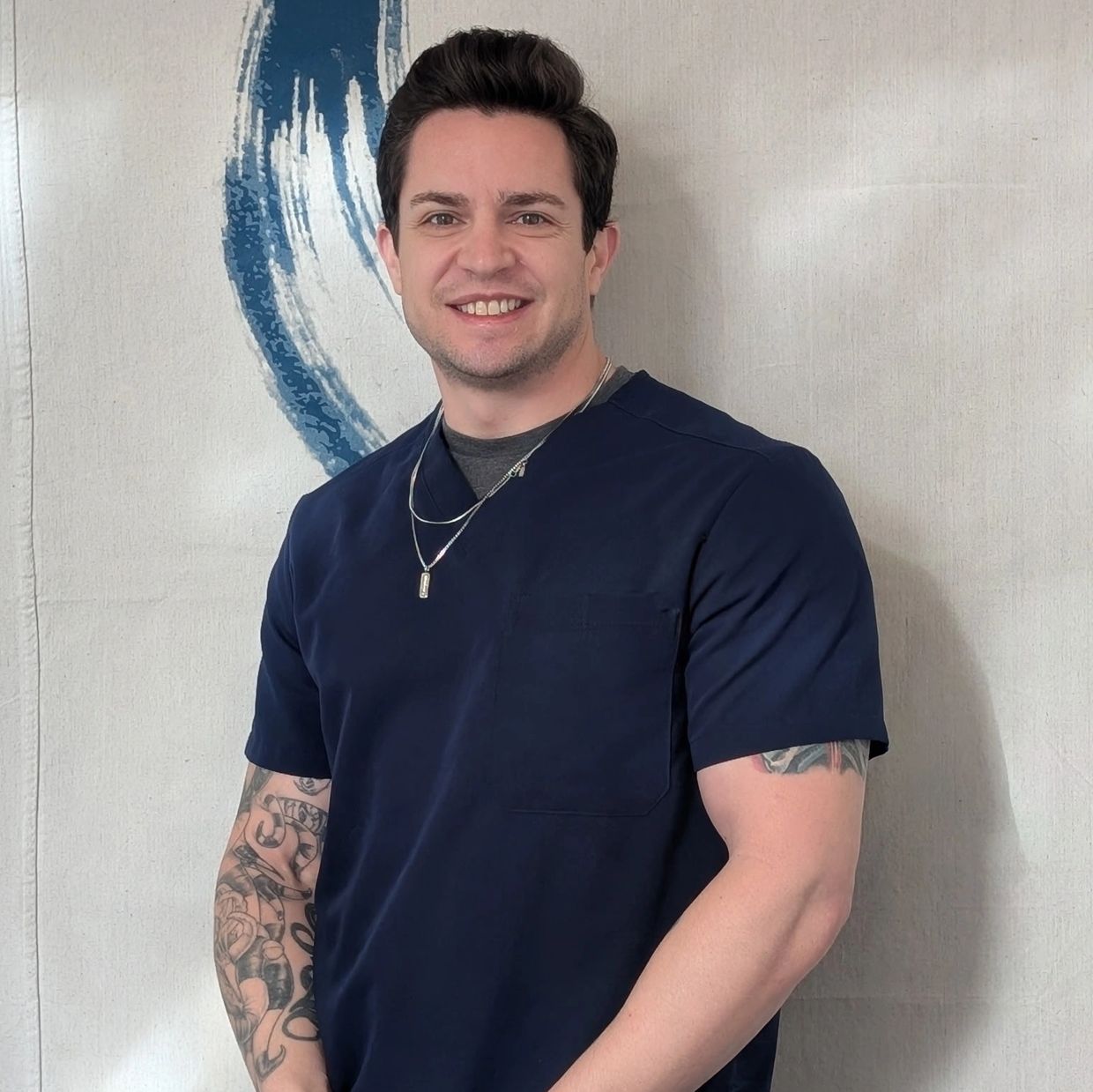 Smiling man with tattoos wearing a navy scrub top and necklaces.