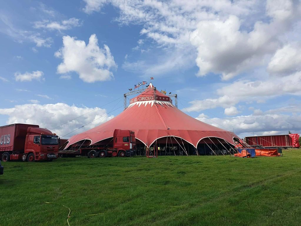 Large red circus tent set up on grass with trucks parked nearby.