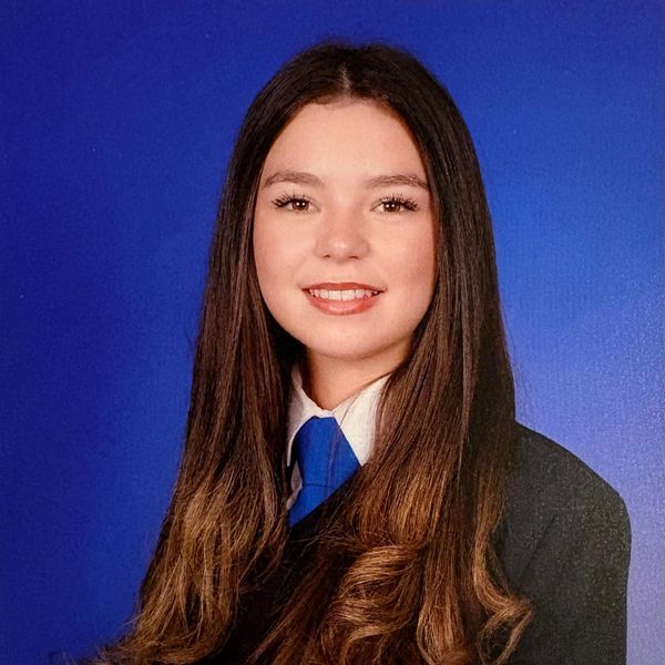 Young woman in formal attire with long brown hair against a blue background.