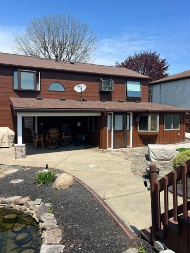 Brown two-story house with a patio and a small pond in the foreground.