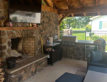 Cozy outdoor patio with stone fireplace, grill, and seating area under wooden roof.