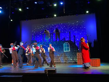 Actors perform a lively scene on stage with a blue starry backdrop.