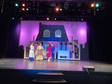 Actors in period costumes on stage with a house backdrop under theatrical lights.
