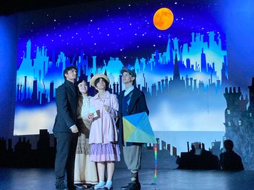 Four actors on stage with a cityscape backdrop and orange moon.