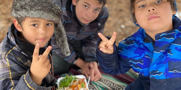 Three boys outdoors showing peace signs with a plate of food on a blanket.