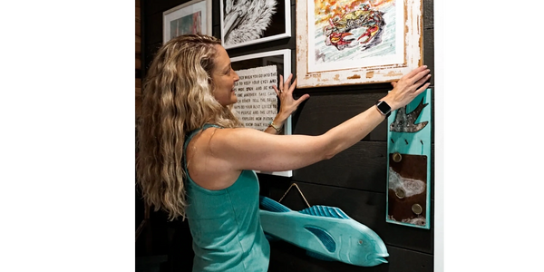 Woman arranging framed artwork on a dark wall with a wooden fish decoration. She is interior stying.