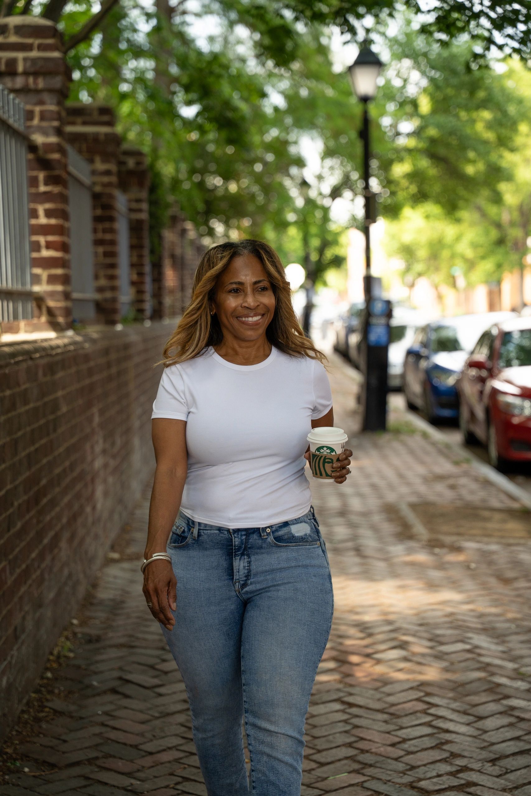 Smiling woman walking down a tree-lined street holding a Starbucks coffee.