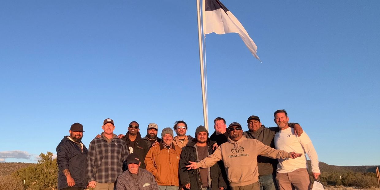 Group of men posing outdoors under a clear blue sky with a flagpole.