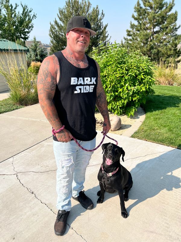 Man with tattoos holding a black dog on a leash outdoors.