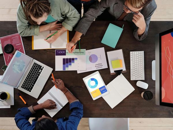 Three people collaborate over charts, laptops, and notes at a wooden table.