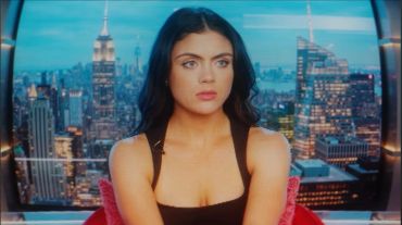 Young woman with dark hair sitting against a cityscape background.