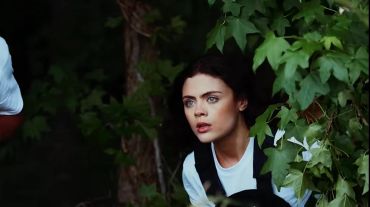 Woman cautiously peering through dense green foliage.